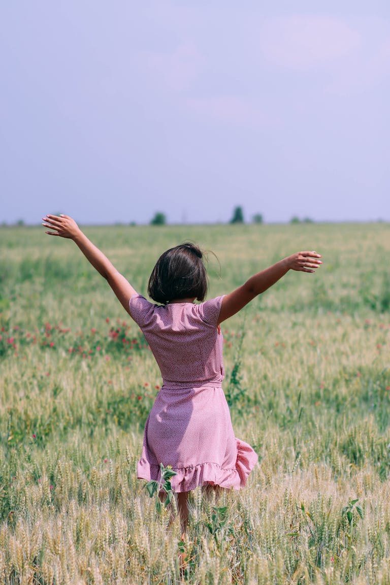 A woman in a pink dress with arms raised in a lush green wheat field under a clear sky.