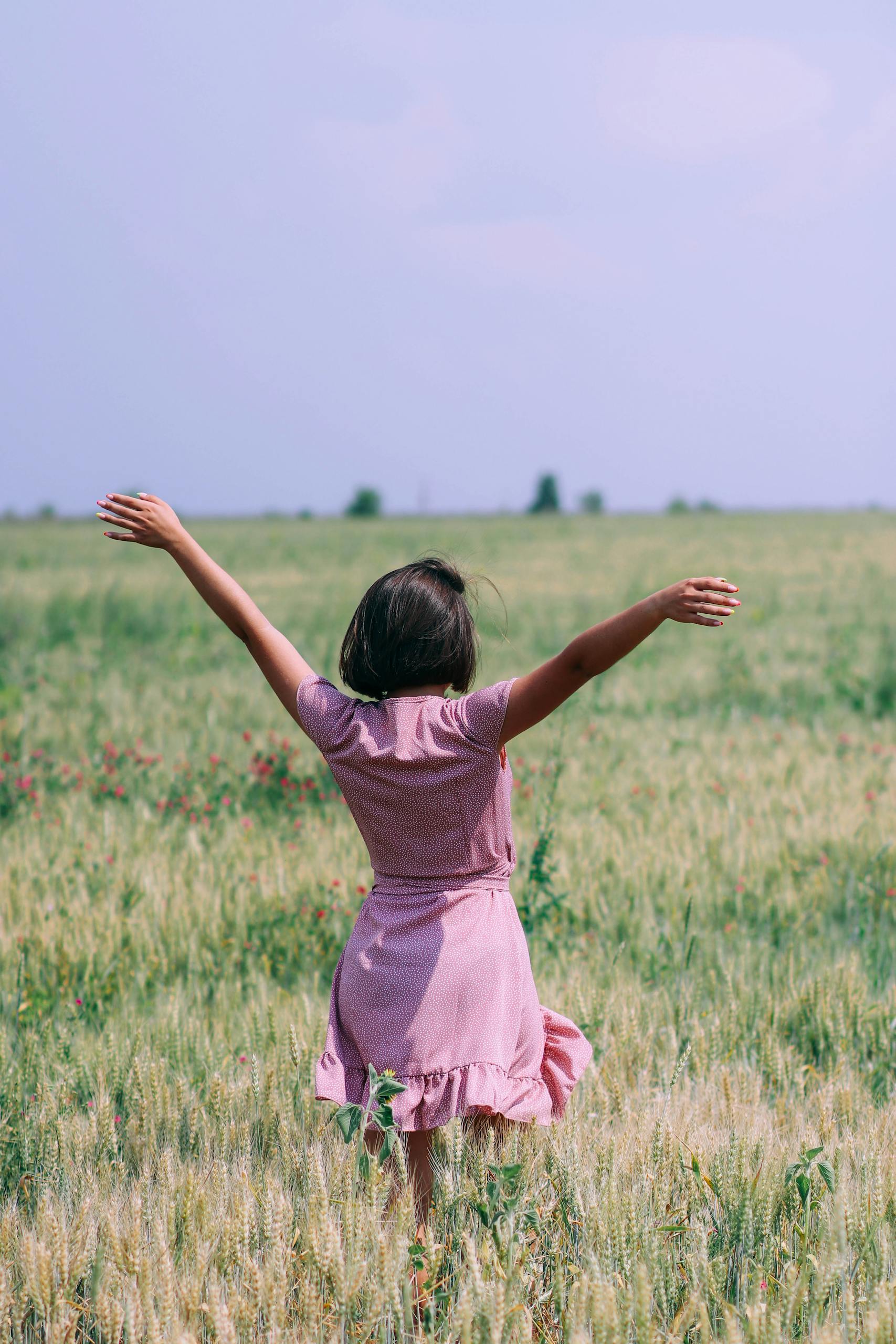 A woman in a pink dress with arms raised in a lush green wheat field under a clear sky.