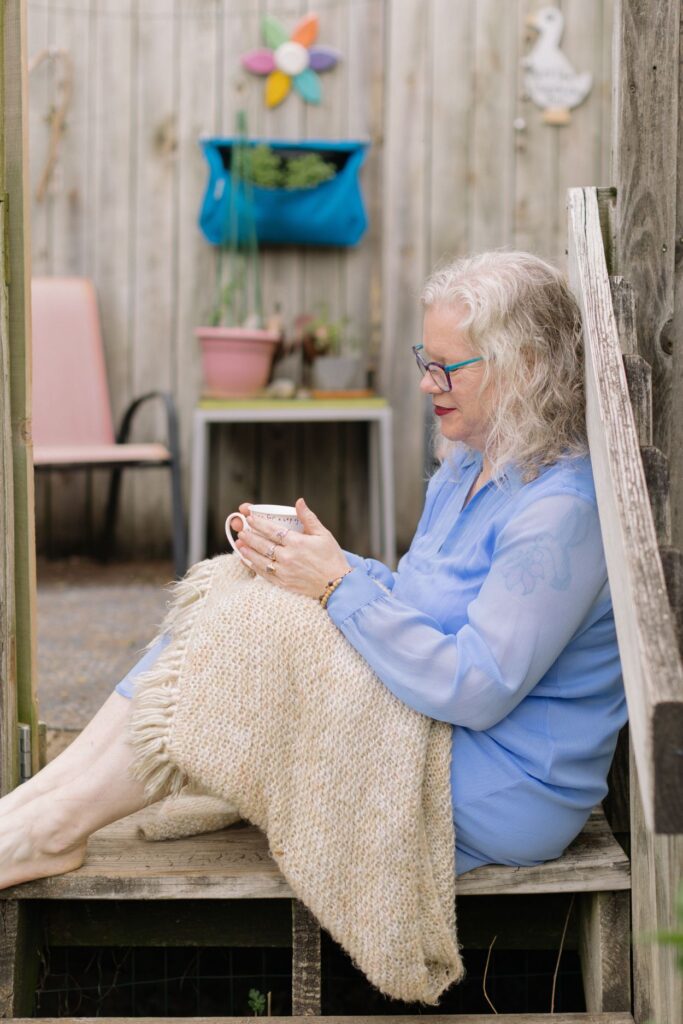 Erika Hall sitting on deck steps with shawl, holding a cup of tea, listening
